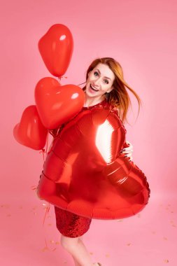 Kırmızı kalpli balon pozu veren kızıl saçlı güzel bir kız. Sevgililer günün kutlu olsun. Pembe arka planda dans eden güzel kızıl kızın stüdyo fotoğrafı..