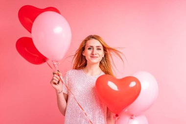 Kırmızı kalpli balon pozu veren kızıl saçlı güzel bir kız. Sevgililer günün kutlu olsun. Pembe arka planda dans eden güzel kızıl kızın stüdyo fotoğrafı..