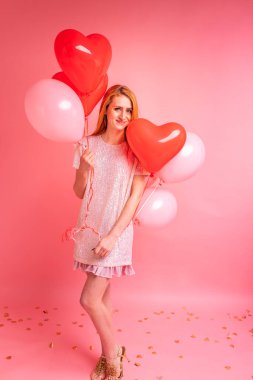 Kırmızı kalpli balon pozu veren kızıl saçlı güzel bir kız. Sevgililer günün kutlu olsun. Pembe arka planda dans eden güzel kızıl kızın stüdyo fotoğrafı..