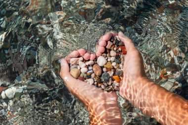 Elleri renkli çakıl taşlarıyla dolu. Deniz dalgalarının arka planında kalp şekli oluşturuyor. Tatil, dinlenme, deniz kenarında tatil