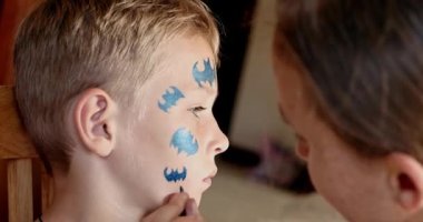 Child animator, artist's hand draws face painting to little boy. Child with funny face painting. Painter makes blue bat mask on boy's face. Face painting at children holiday, event, birthday party, entertainment.