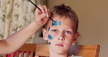 Child animator, artist's hand draws face painting to little boy. Child with funny face painting. Painter makes blue bat mask on boy's face. Face painting at children holiday, event, birthday party, entertainment.