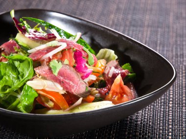 Biftek dilimli salata, yeşil marul, salatalık, domates ve soğan siyah hasır bir tabak hasırda.