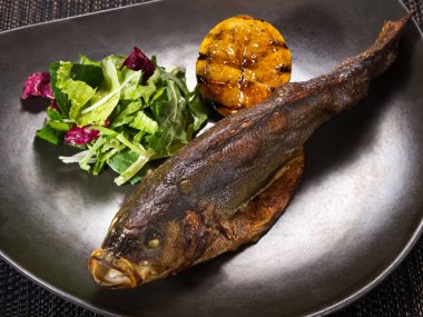 Koyu arkaplanda ızgara limon ve salata karışımı tabakta pişmiş alabalık.