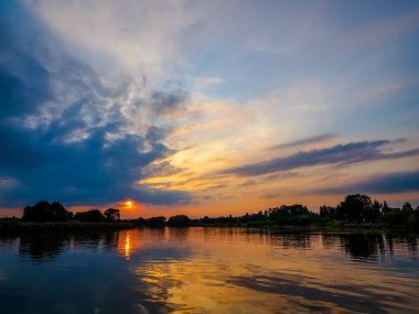 Akşamları nehir üzerinde dramatik bulutlarla gökyüzü. Bulutlar ve nehirde dramatik gün batımı. Doğa manzarası, yansıma, mavi bulutlu gökyüzü ve turuncu güneş ışığı, gün batımında manzara. 