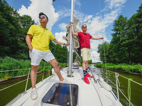 两个英俊的年轻人穿着休闲装 站在帆船船头上 望着前方阳光灿烂的夏日 在游艇上放暑假 — 图库照片