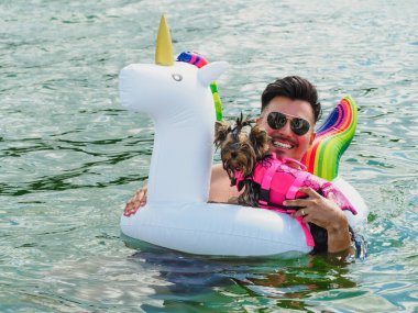 Gülümseyen yakışıklı bir genç Yorkshire teriyeriyle yüzer suya can yeleği giyer.