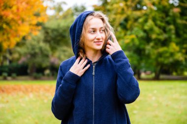 Güz mevsiminde, kafasında uzun kapüşonlu kapüşonlu güzel sarışın kadın şehir parkının dış tarafına bakıyor.