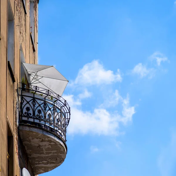 Old balcony with open umbrella on a facade of building on a blue sky and clouds background. square photo