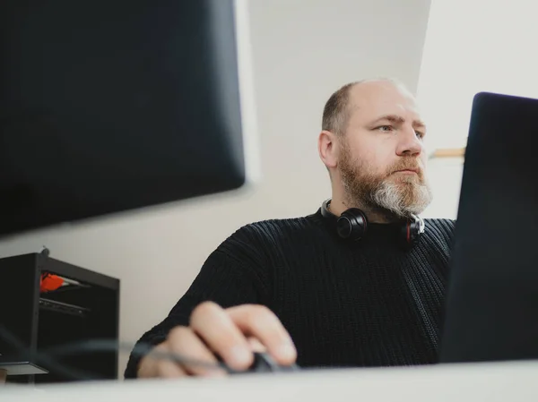Adult focused bearded man remote working on laptop and using two monitors from home
