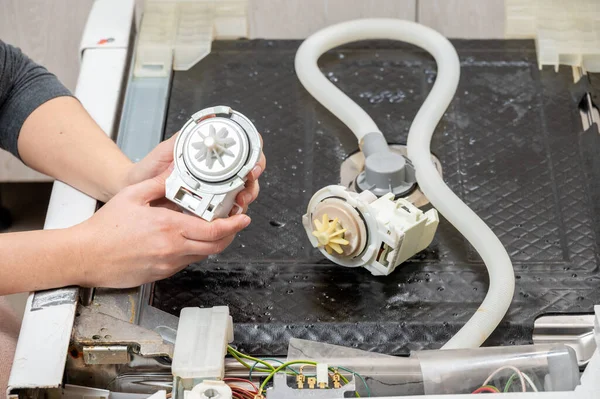 A new pump for a dishwasher in the hands of a woman, against the background of a disassembled dishwasher