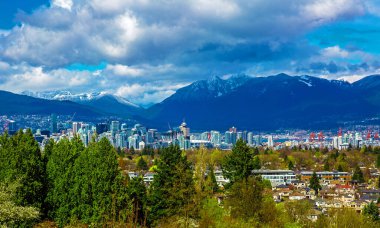 Vancouver şehir merkezi ve liman manzarası Rocky Dağları sıradağları ve arka planda fırtınalı gökyüzü ve ön planda yeşil ağaçlarla şehir manzarası