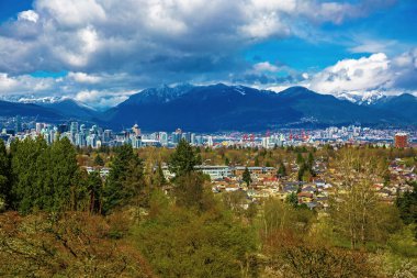 Vancouver şehir merkezi ve liman manzarası Rocky Dağları sıradağları ve arka planda fırtınalı gökyüzü ve ön planda yeşil ağaçlarla şehir manzarası
