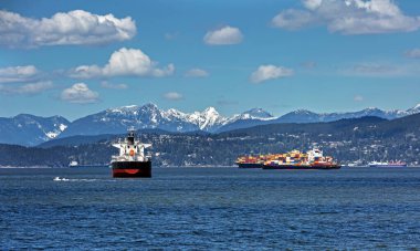 Vancouver Limanı 'ndaki okyanus gemileri ve konteyner gemileri güneşli bir günde karla kaplı bir dağ sırtı ve bulutlu bir gökyüzüne karşı