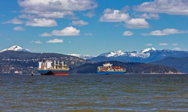 Vancouver Limanı 'ndaki okyanus gemileri ve konteyner gemileri güneşli bir günde karla kaplı bir dağ sırtı ve bulutlu bir gökyüzüne karşı