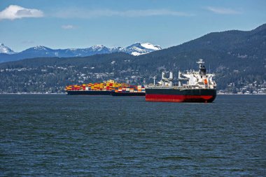 Vancouver Limanı 'ndaki okyanus gemileri ve konteyner gemileri güneşli bir günde karla kaplı bir dağ sırtı ve bulutlu bir gökyüzüne karşı