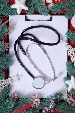 Tıbbi steteskoplu pano, kırmızı arka planda köknar dalları ve noel süslemeleri çerçeveli. 