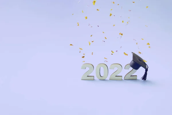 Mezuniyet 2022, ahşap bir 2022 sayısında, uçan konfetiyle çok renkli bir arka planda mezuniyet şapkası takıyor.