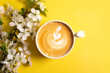 Sarı arka planda çiçek açan kiraz dallı seramik bir fincanda kahve sanatıyla kapuçino manzarası..