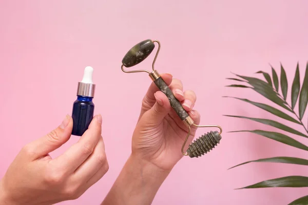 jade stone roller massager and cosmetic dropper bottle in female hands. SPA and beauty therapy. Pink background with palm leaf decoration.