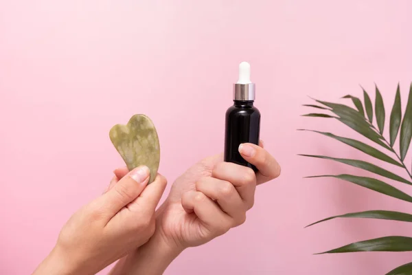 jade stone massager gua sha and cosmetic dropper bottle in female hands. SPA and beauty therapy. Pink background with palm leaf decoration.