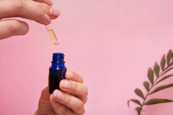 Cosmetic oil in dropper bottle in female hands. SPA and beauty therapy. Pink background with palm leaf decoration.