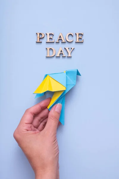 Peace day text from wooden letters with yellow-blue paper dove in female hand on blue background top view. International Peace day concept. Vertical format