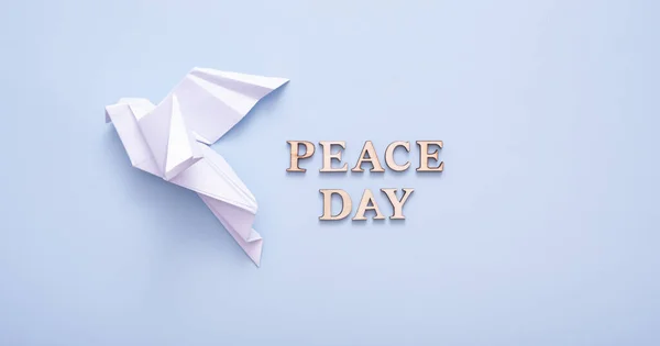 Peace day text from wooden letters with white paper dove in female hand on blue background. International Peace day concept. Vertical photo.