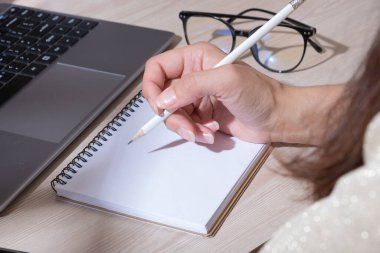 Cropped shot of woman hand writing on notebook with pencil while working with computer at her office desk.