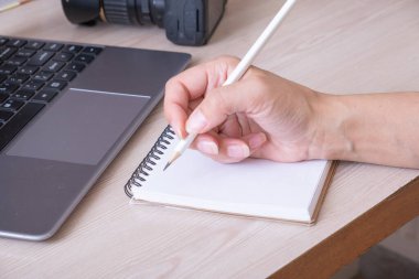 Notebooks and female hand writes with a pencil near a laptop and a photocamera at a desk.