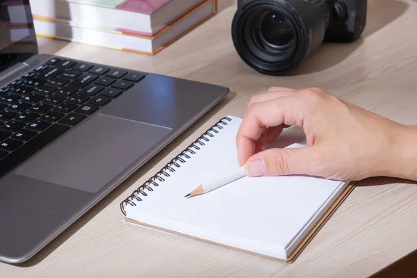 Female hand with a pencil on notebook near a laptop and a camera and books at a desk.