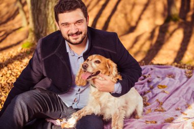 Paltolu genç adam, sonbahar ormanında köpekle birlikte arka planda renkli yapraklarla resmedilmiş.