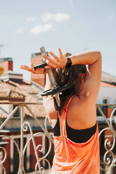 Вписується жінка в чорно-помаранчевий спортивний одяг, вправляється з гантелями на даху в сонячний день. Концепція фітнесу — стокове фото