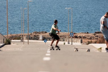 Güneşli bir günde kırsal bir yolda artistik patinaj yapan iki adam sahilde sörf yapıyor.