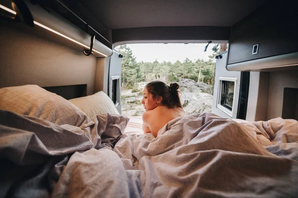 Young nude female lying on bed under blanket and looking away over shoulder against picturesque coniferous forest behind opened camper doors during road trip in summer