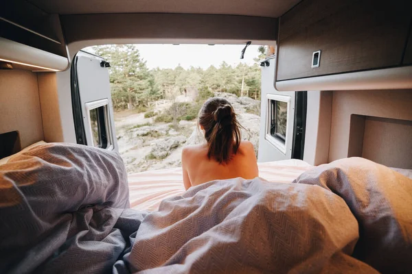 Young nude female lying on bed under blanket and looking away over shoulder against picturesque coniferous forest behind opened camper doors during road trip in summer
