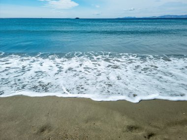 Deniz dalgalı su köpüğü ıslak kumda, mavi gökyüzü arka planında. Boş kumlu plaj, Yunan adası Cyclades, Yunanistan. Yaz tatili şablonu, boşluk.