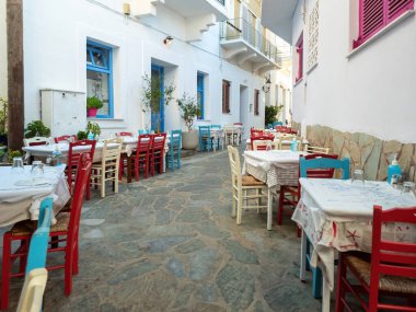 Andros Adası Cyclades Yunanistan 'ın Batsi köyündeki Yunan açık hava tavernası restoranı. Parke taşına boş masa ve sandalyeler. Binalar, fabrikalar.