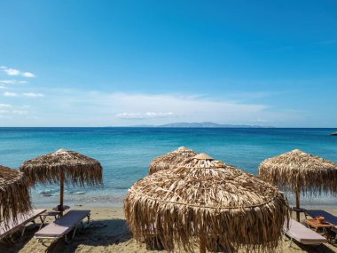 Yunanistan. Yunan yaz güneşinin altında boş şemsiyeler. Sandy Kalivia Plajı, Tinos Adası, Cyclades. Mavi gökyüzü sakin deniz arka planı.