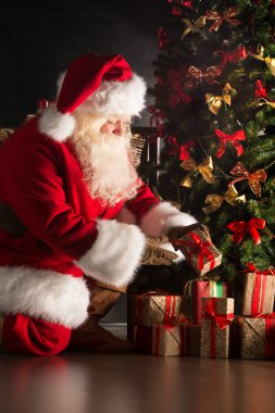 Santa putting gifts under Christmas tree in dark room clipart