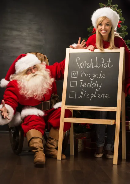 Papai Noel com seu ajudante de pé perto quadro com lista de desejos — Fotografia de Stock