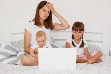 Evde küçük çocuklarla çalışmak, dizüstü bilgisayarla uğraşan genç yetişkin bir anne, çocuklarıyla yatakta otururken birden fazla iş yapmaya çalışan stresli bir anne..
