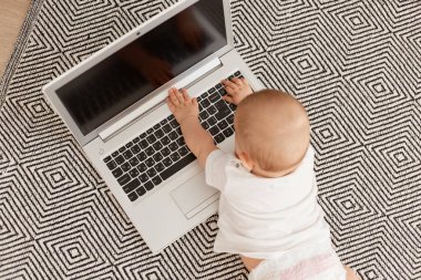 Beyaz tişörtlü bebek, gri halıda yerde yatarken, dizüstü bilgisayarlı, boş ekranlı, küçük bebek bilgisayar kullanıyor..