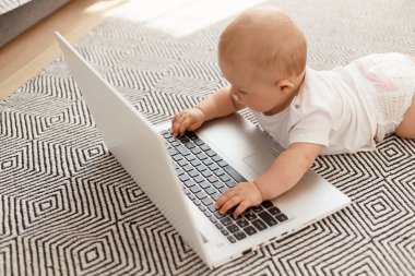 Sevimli bebek modern teknolojiyle ilgileniyor, yere uzanıyor ve dizüstü bilgisayar kullanıyor annesinin neden gittiğini, küçük kız gri halıda, elinde defter var..