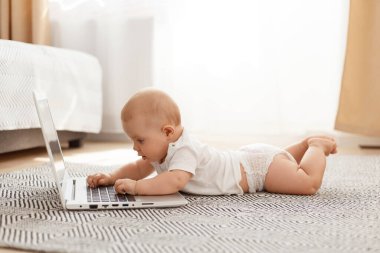 Beyaz tişörtlü, gri halıda yerde yatıp dizüstü bilgisayar okuyan ya da çizgi film izleyen küçük bebek portresi..