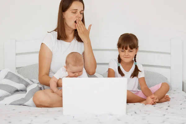 疲惫困倦的妇女母亲与孩子坐在床上 用笔记本电脑工作 打呵欠 用手掌捂住嘴 任务繁多 自由职业和做母亲的形象 — 图库照片