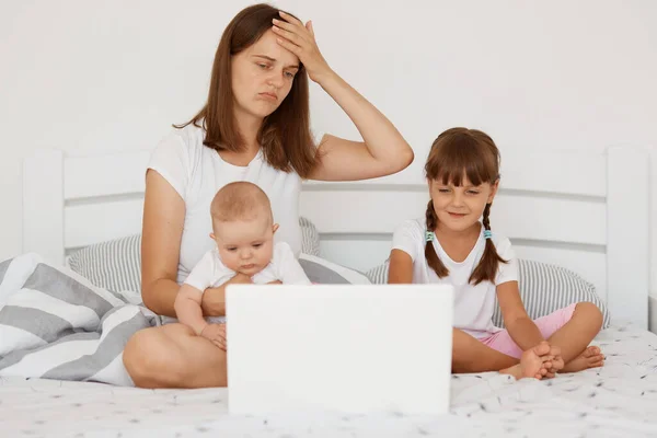 子守をしながらノートパソコンで仕事をしようとする若い大人の母親ストレスの多いお母さん子供たちとベッドに座ってマルチタスクをしようとする — ストック写真