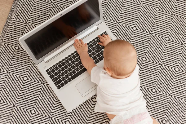 Beyaz tişörtlü bebek, gri halıda yerde yatarken, dizüstü bilgisayarlı, boş ekranlı, küçük bebek bilgisayar kullanıyor..