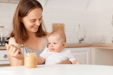 Beyaz tenli, siyah saçlı bir kadın küçük sevimli kızını meyve ve sebze püresiyle besliyor. İnsanlar ışık odasında poz veriyor, sevgi ve özen gösteriyorlar..