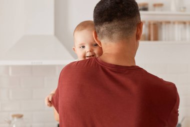 Koyu renk saçlı, sıradan bir tişört giyen, kameraya ters poz veren, küçük kızına ya da oğluna sarılan, arka planda mutfak setiyle evde poz veren bir adam..